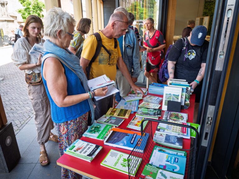 Een groep mensen staat voor het Duurzaamheidscentrum. Rechts is een rode tafel met daarop verschillende boeken met duurzame onderwerpen. Voor de tafel staan verschillende bezoekers die door boeken bladeren.